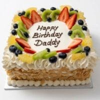 Egg-free fresh fruit celebration cake topped with strawberries, kiwi, blueberries, orange segments, cream piping and almond flakes with a personalised birthday message.