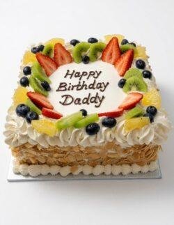 Egg-free fresh fruit celebration cake topped with strawberries, kiwi, blueberries, orange segments, cream piping and almond flakes with a personalised birthday message.