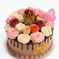 Round chocolate fresh cream cake decorated with chocolate drip, handcrafted fresh cream roses in pink, ivory, burgundy, peach and orange, a golden crown topper, and butterfly decorations.