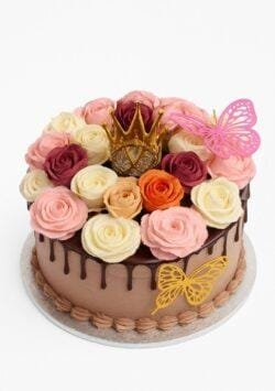 Round chocolate fresh cream cake decorated with chocolate drip, handcrafted fresh cream roses in pink, ivory, burgundy, peach and orange, a golden crown topper, and butterfly decorations.