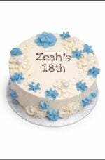 Tall white fresh cream birthday cake decorated with blue and white buttercream flowers, edible pearl accents, and a personalised birthday message.