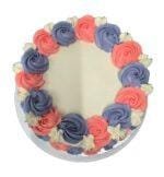 Top view of a white fresh cream celebration cake featuring pink and lavender fresh cream rosettes arranged around the edge with white floral accents and a personalised message area.