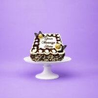 Square fresh cream celebration cake decorated with chocolate and vanilla swirls, white floral piping, Ferrero Rocher chocolates, chocolate leaf decorations, milk and white chocolate shavings, and a personalised message.