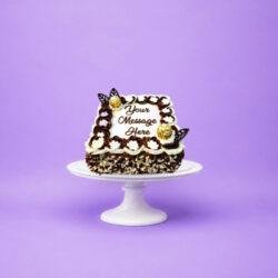 Square fresh cream celebration cake decorated with chocolate and vanilla swirls, white floral piping, Ferrero Rocher chocolates, chocolate leaf decorations, milk and white chocolate shavings, and a personalised message.